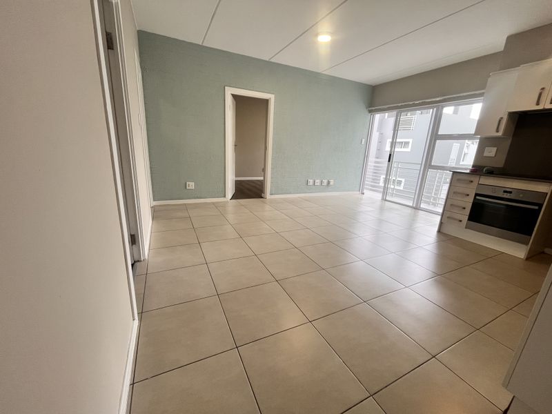 An open-plan living and kitchen area featuring neutral tiled flooring, modern cabinetry with a built-in oven, and access to a balcony through sliding glass doors.