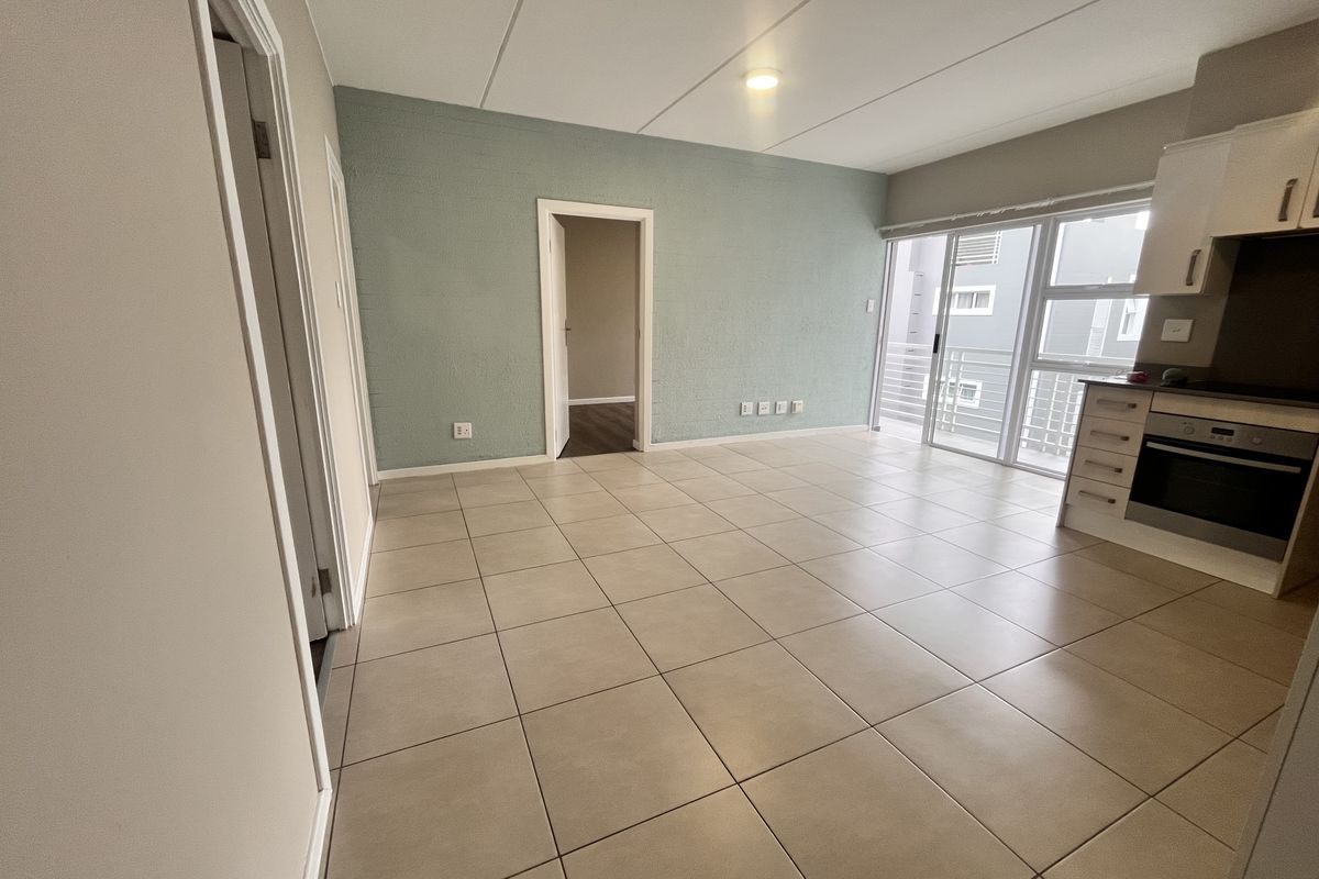An open-plan living and kitchen area featuring neutral tiled flooring, built-in kitchen cabinetry with integrated oven, and access to a balcony through sliding doors.