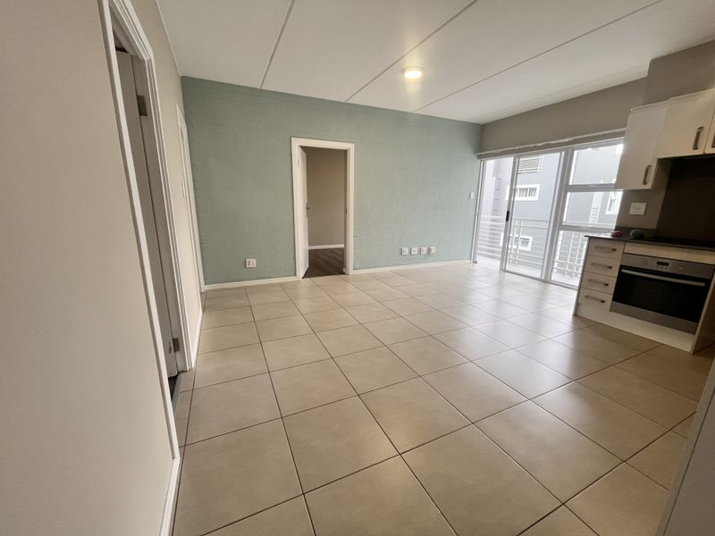 An open-plan living and kitchen area featuring neutral tiled flooring, built-in kitchen cabinetry with integrated oven, and access to a balcony through sliding doors.