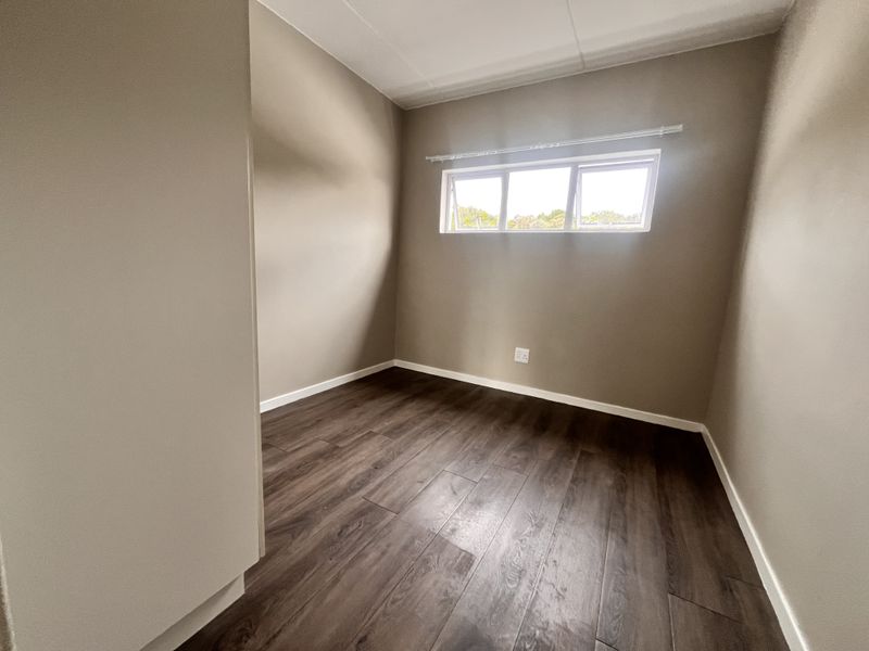A clean, empty bedroom featuring modern dark laminate flooring, neutral wall paint, and a top-hung window providing natural light.