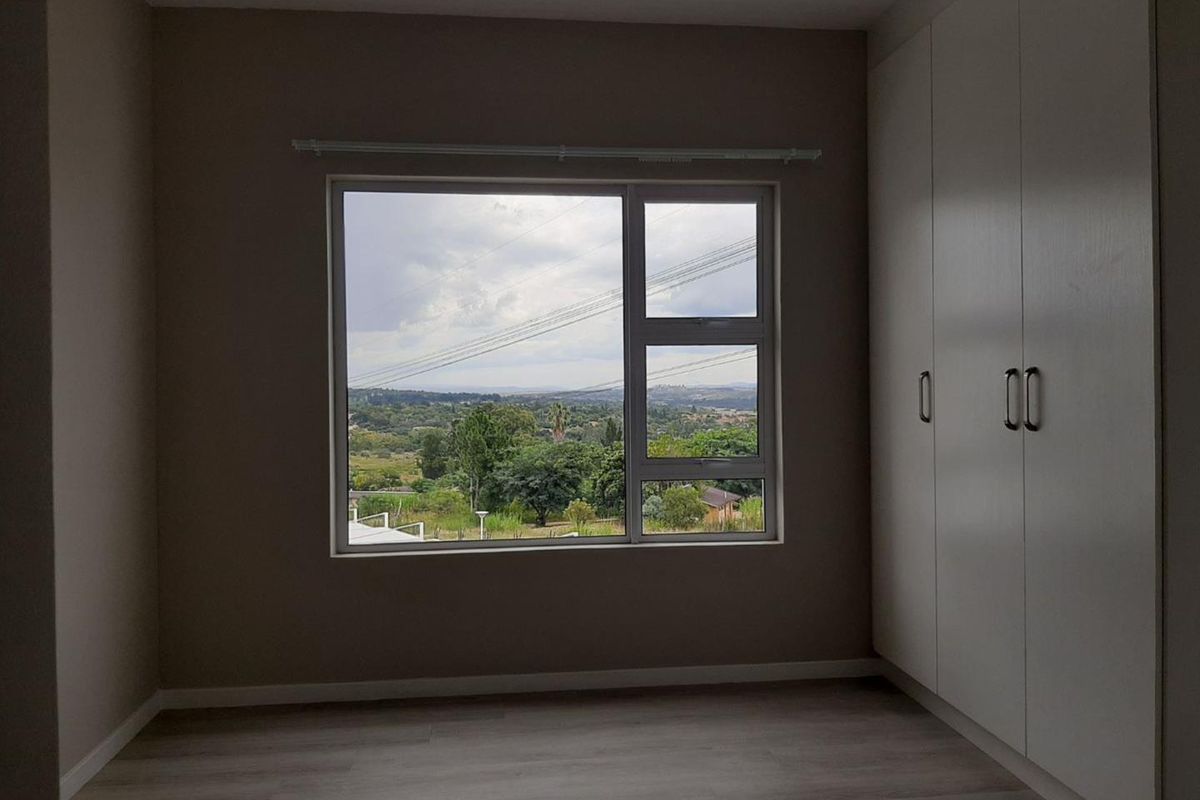 A bright, empty bedroom featuring modern laminate flooring, built-in cupboards, and a large window offering scenic landscape views.