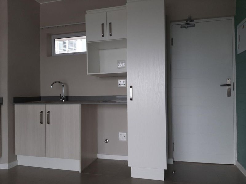 A compact, modern kitchen area featuring light wood-finish cabinetry, a stainless steel sink, and a tall pantry cupboard, presented in a clean, empty space.