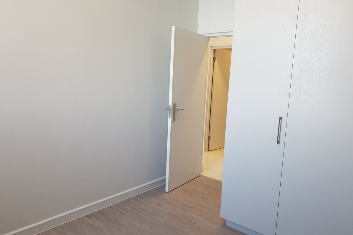 A clean, empty bedroom featuring modern laminate flooring, neutral walls, and built-in cupboards with a view into the adjacent hallway.