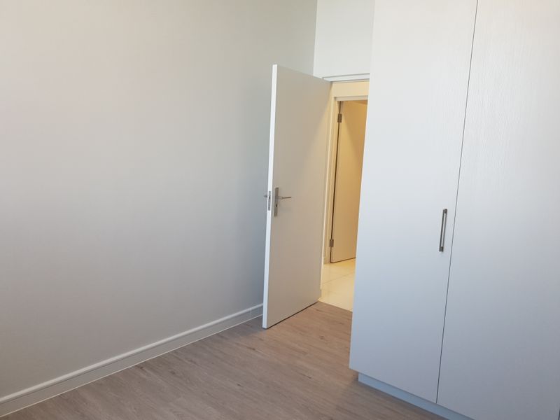 A clean, empty bedroom featuring modern laminate flooring, neutral walls, and built-in cupboards with a view into the adjacent hallway.
