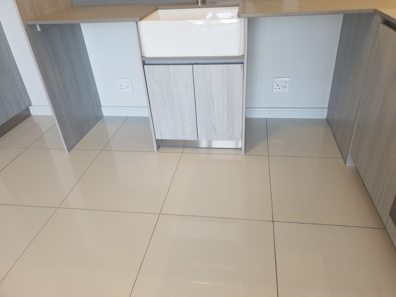 A modern kitchen area featuring a white farmhouse sink, light wood-grain cabinetry, stone countertops, and polished floor tiles.