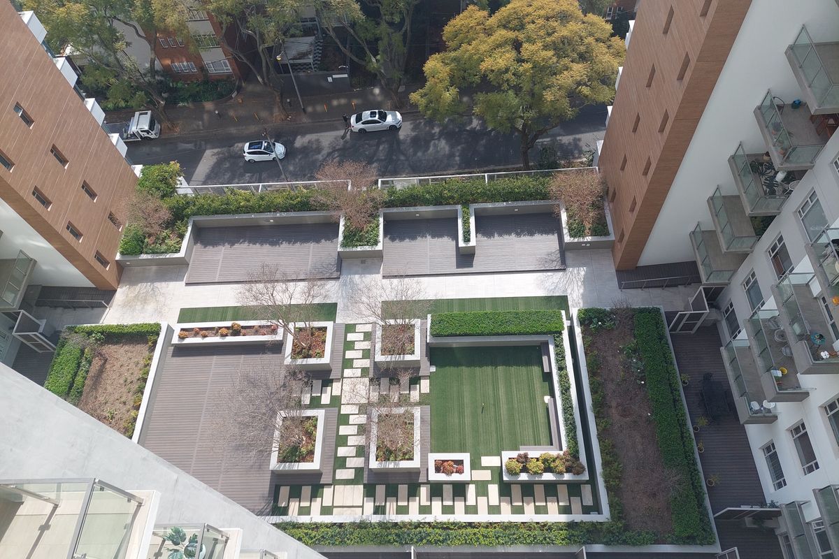 An elevated aerial view of a modern apartment complex courtyard featuring landscaped gardens, paved walkways, and wooden decking areas.