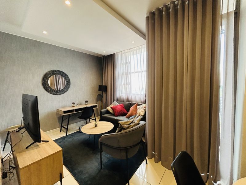 A well-maintained living room featuring modern recessed ceiling lighting, textured feature wall, and large windows with floor-to-ceiling curtains, finished with neutral tiled flooring.