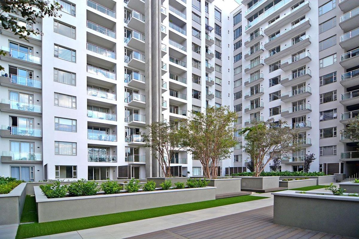 A view of the apartment complex courtyard featuring landscaped garden beds, paved walkways, and modern building facades with private balconies.