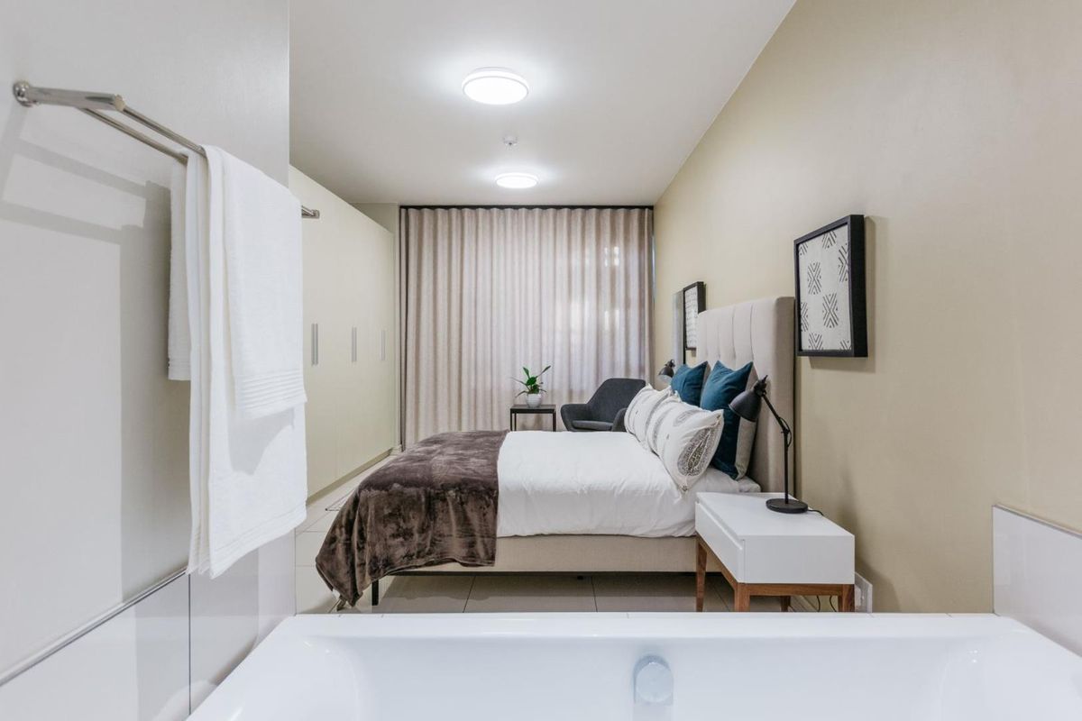 A modern bedroom featuring built-in wardrobes, neutral wall finishes, and an integrated bathroom layout with a visible bathtub in the foreground.