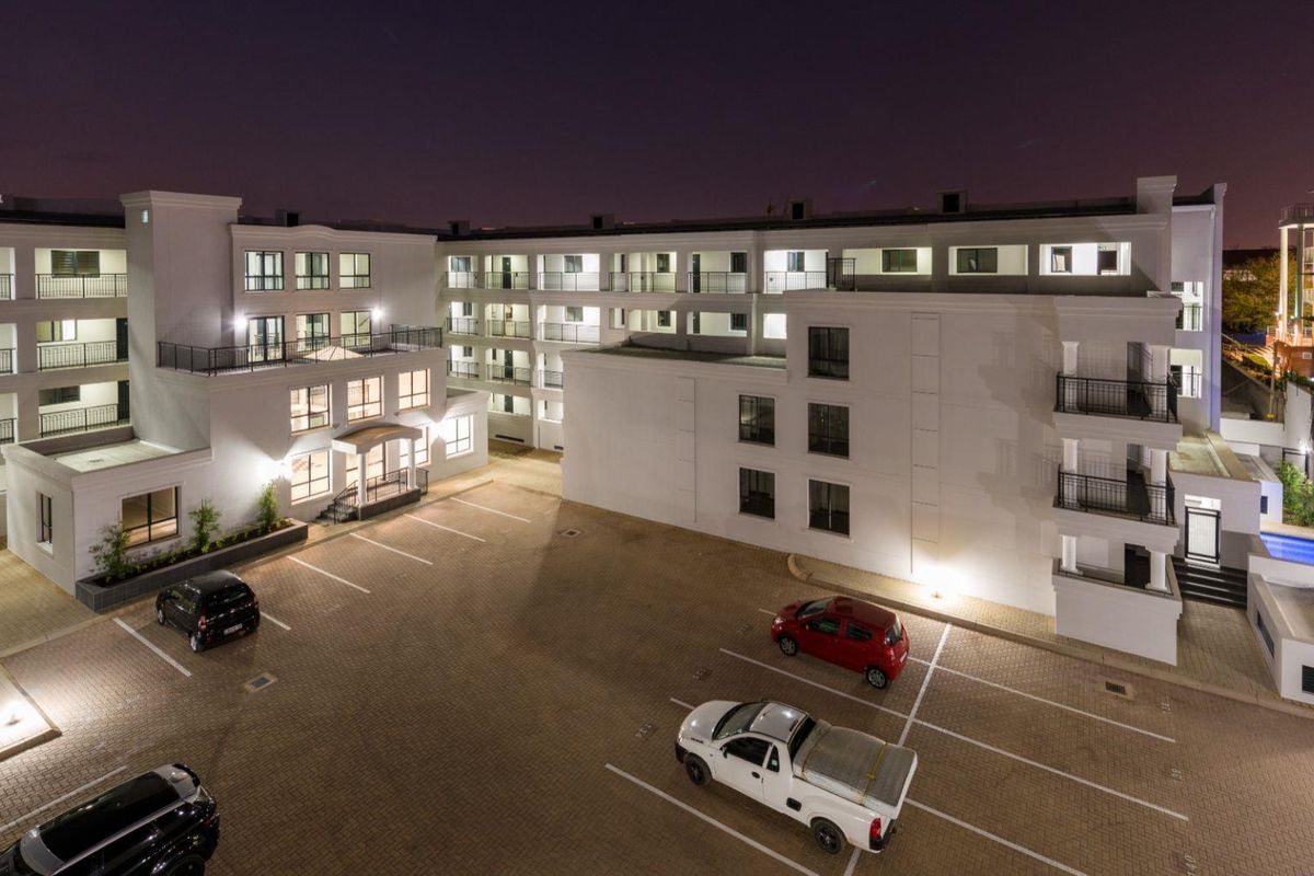 An elevated night view of the apartment complex exterior, featuring paved parking areas, multi-story building facades, and balcony access.