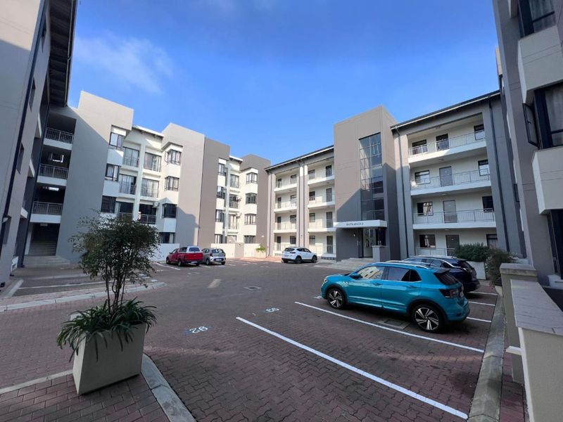 Exterior view of a modern apartment complex featuring a paved parking area, multi-story residential blocks with balconies, and a designated building entrance.