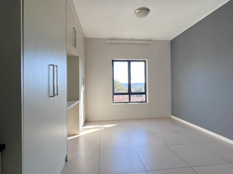 A bright, empty bedroom featuring neutral tiled flooring, built-in cupboards, and a large window with natural light.