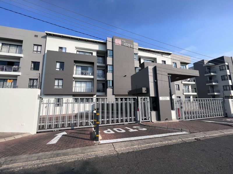Front exterior view of The Atrium apartment complex featuring a secure gated entrance, paved driveway, and modern multi-story building facade with balconies.