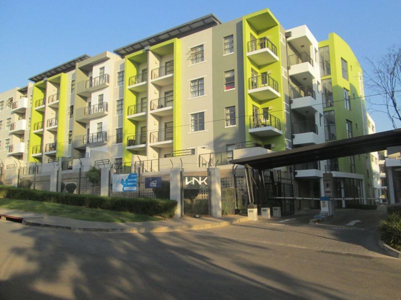 Modern apartment complex exterior featuring green and grey facade, private balconies, secure gated entrance, and paved driveway access.