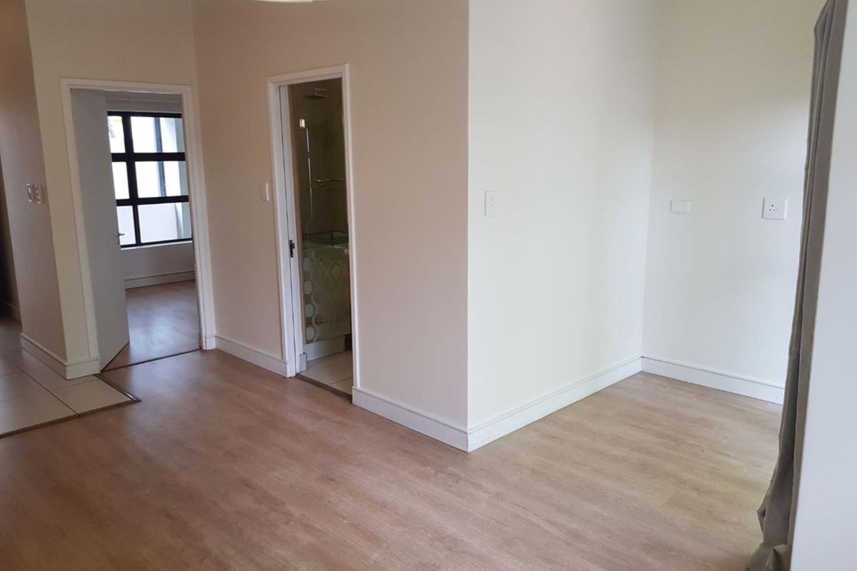 An empty living area featuring light-toned laminate flooring, neutral walls, and access to adjacent rooms including a bathroom.