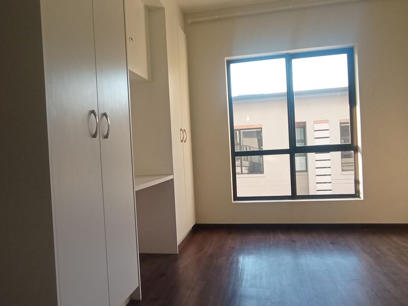 A bright, empty bedroom featuring modern laminate flooring, built-in cupboards, and a convenient integrated desk space.