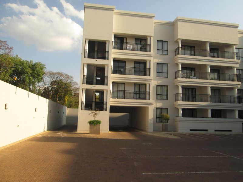 Front view of a multi-story apartment complex featuring paved driveway access, covered parking area, and private balconies with metal railings.