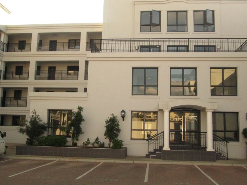 Front elevation of a multi-story apartment building featuring a paved parking area, structured entrance with columns, and private balconies with metal railings.