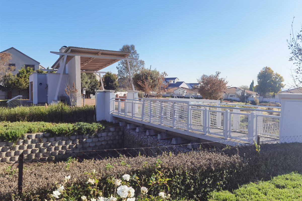 A well-maintained pedestrian bridge with metal railings leading to a covered outdoor entertainment area within a residential estate setting.