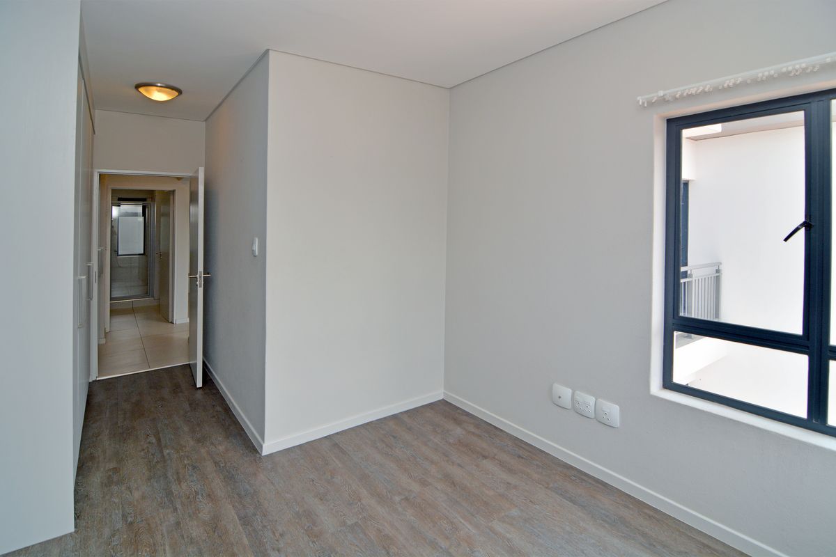 A bright, empty bedroom featuring modern laminate flooring, built-in cupboards, and a window overlooking the building exterior.