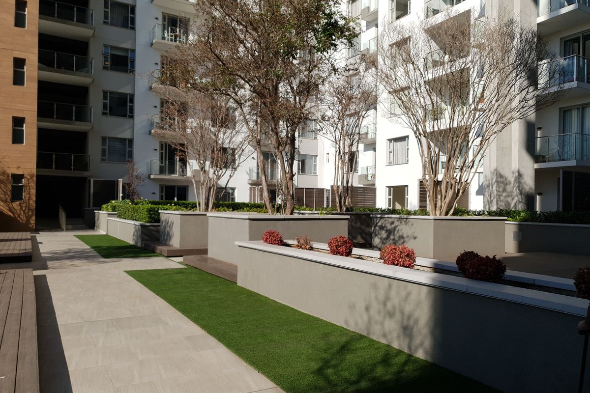 A well-maintained outdoor courtyard area featuring paved walkways, artificial grass sections, and raised planter beds with established trees and shrubs.
