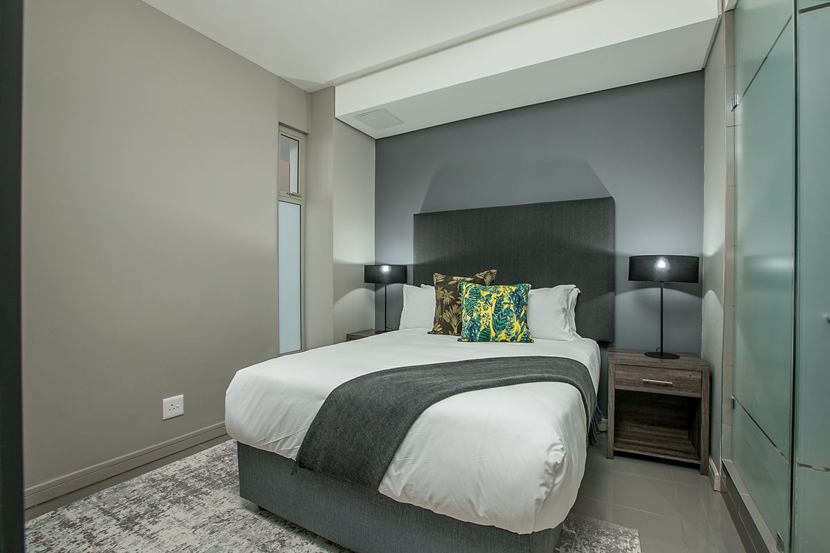A modern bedroom featuring a built-in headboard, recessed ceiling detail, and an adjacent glass-enclosed bathroom area.