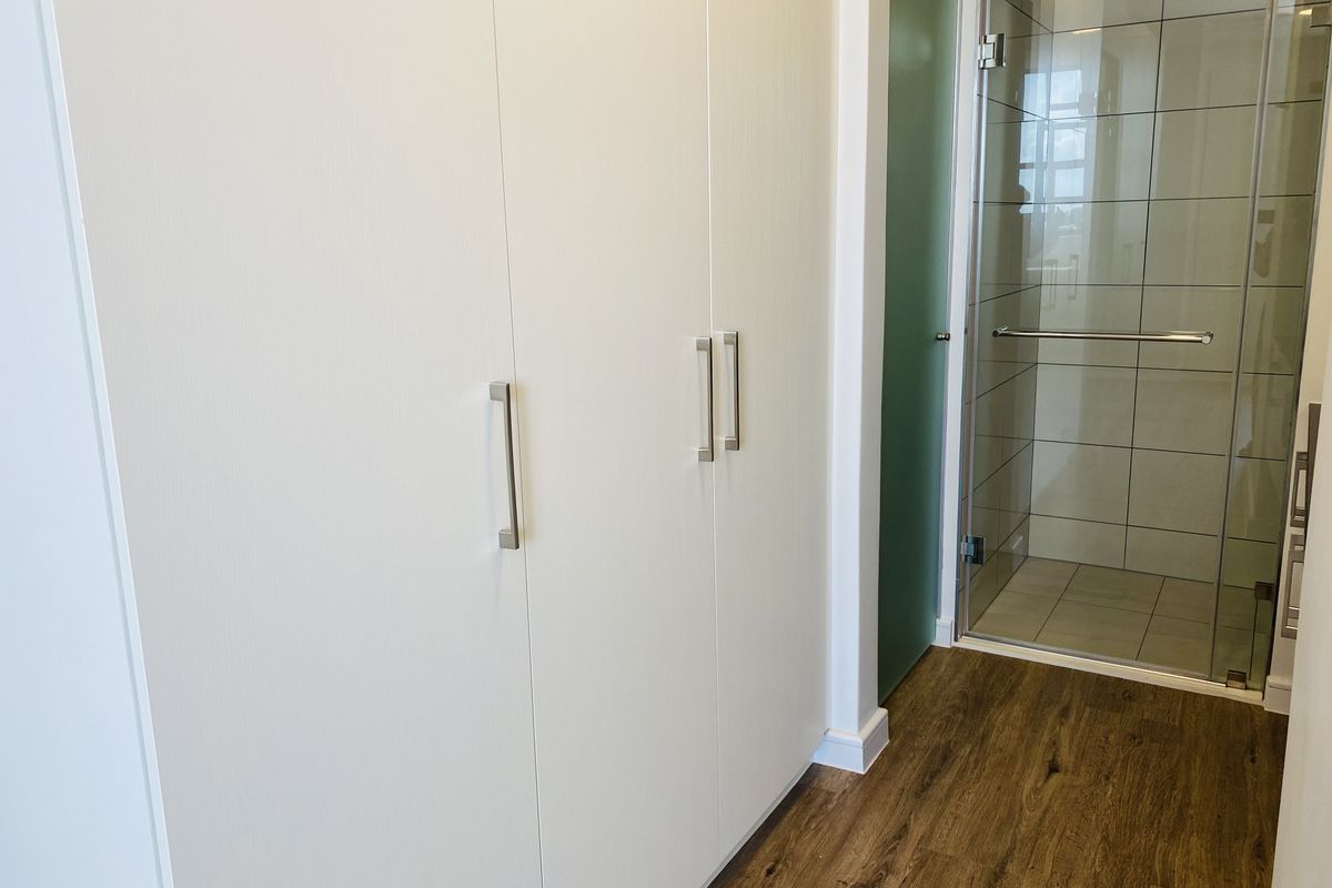 A clean, modern bathroom area featuring built-in storage cupboards, laminate flooring, and a glass-enclosed shower with tiled walls.