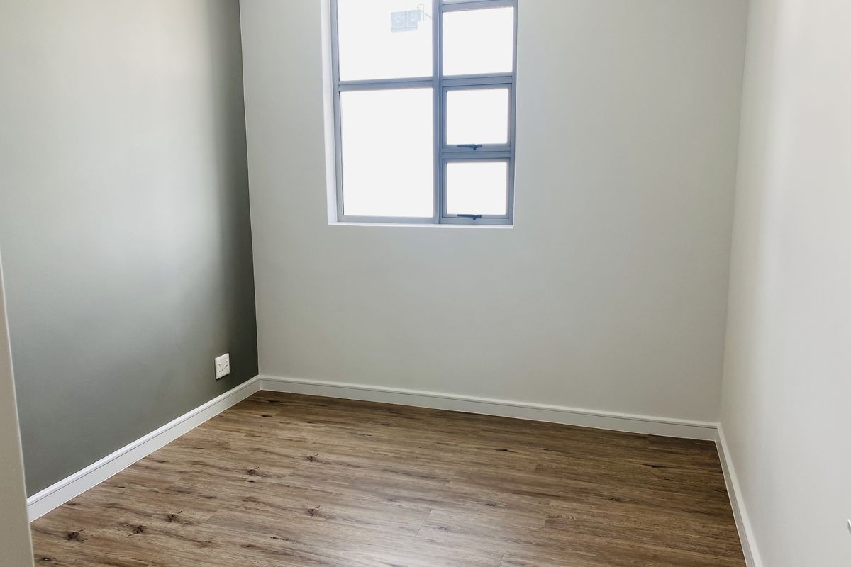 A bright, empty bedroom featuring modern laminate wood-look flooring, neutral painted walls, and a large aluminium-framed window.