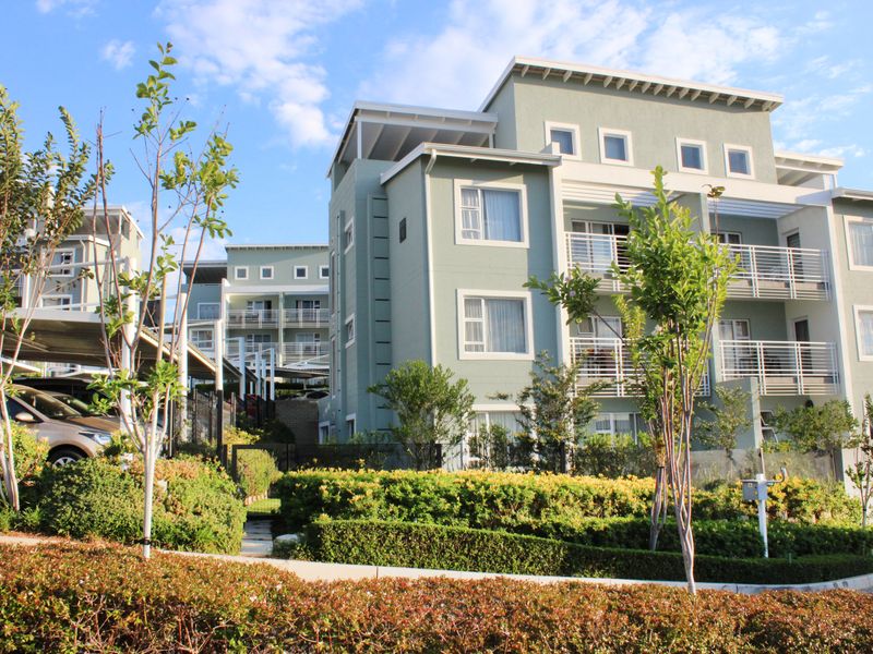 A multi-story apartment complex featuring modern architecture, private balconies with metal railings, a landscaped garden, and covered parking facilities.
