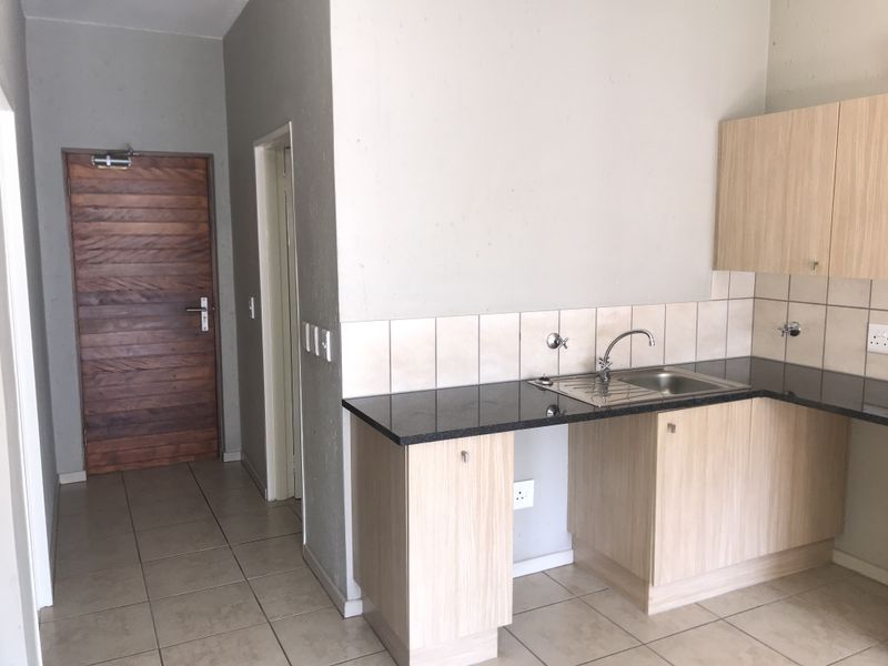 A functional kitchen area featuring light wood cabinetry, granite countertops, a stainless steel sink, and neutral tiled flooring, adjacent to the main entrance.