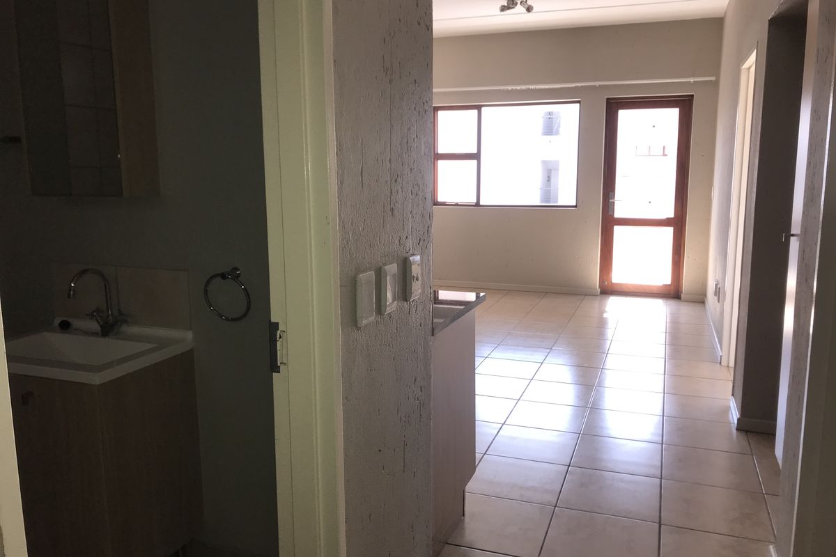 An open-plan apartment interior featuring tiled flooring, a bathroom vanity area, and natural light from the window and door leading to the exterior.