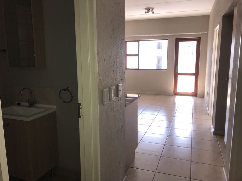 An open-plan apartment interior featuring tiled flooring, a bathroom vanity area, and natural light from the window and door leading to the exterior.
