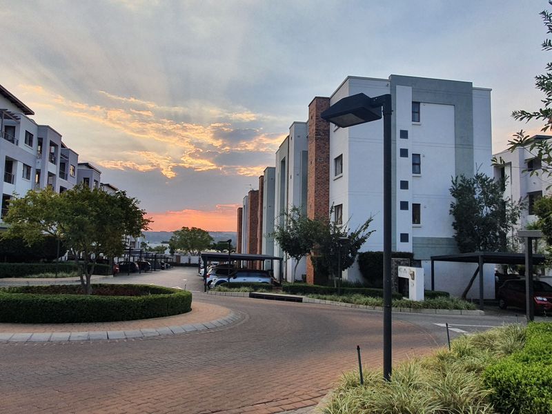 A view of the apartment complex entrance featuring paved driveways, landscaped gardens, and covered parking bays under a sunset sky.