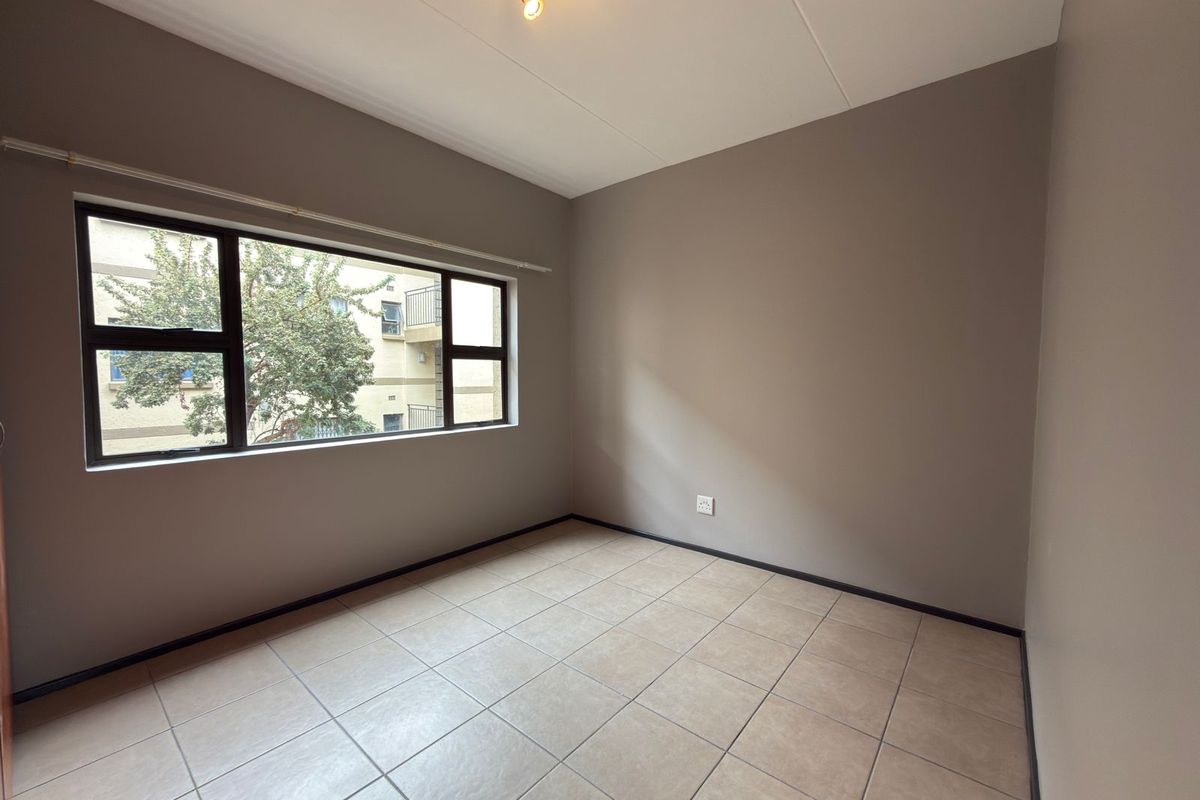 A clean, empty bedroom featuring neutral wall paint, tiled flooring, and a large aluminium-framed window providing natural light.