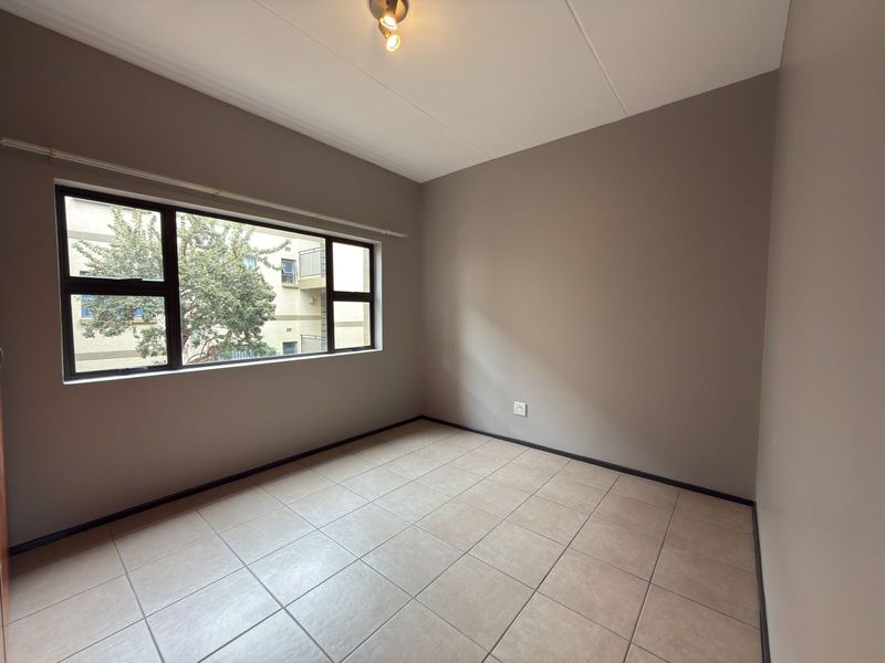 A clean, empty bedroom featuring neutral wall paint, tiled flooring, and a large aluminium-framed window providing natural light.