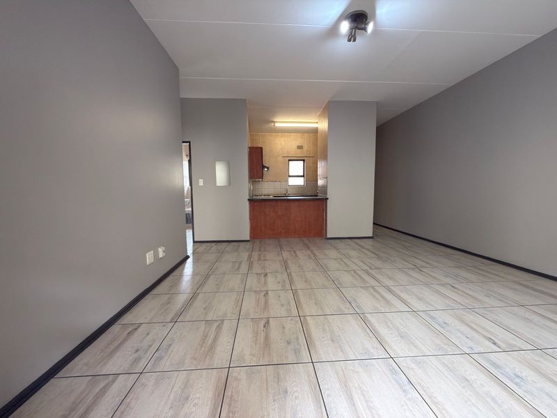 An open-plan living area featuring neutral grey walls, wood-look tiled flooring, and a compact kitchen with built-in cabinetry.
