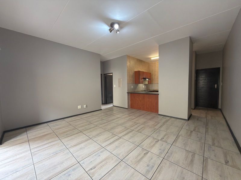 An open-plan living area featuring neutral grey walls, wood-look tiled flooring, and a compact kitchen with built-in cabinetry and an extractor fan.