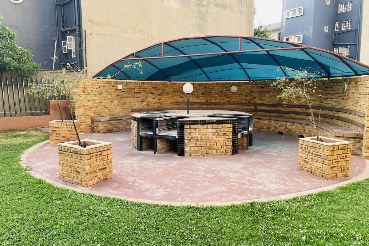 Communal outdoor entertainment area featuring a built-in brick braai, circular seating, and a protective canopy set within a landscaped garden.
