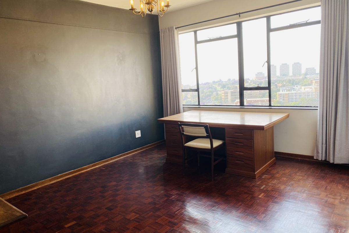 A spacious bedroom featuring classic parquet flooring, a large window with city views, and a dark accent wall.
