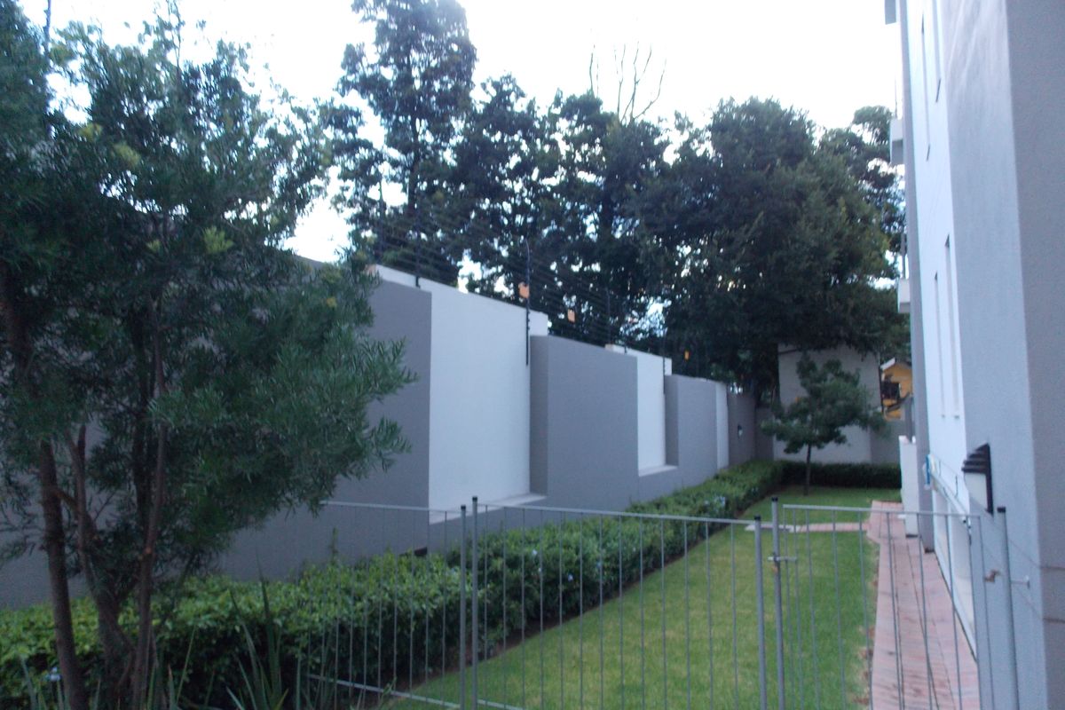 A view of the apartment's rear exterior area, featuring a manicured lawn, perimeter boundary wall with electric fencing, and established garden landscaping.