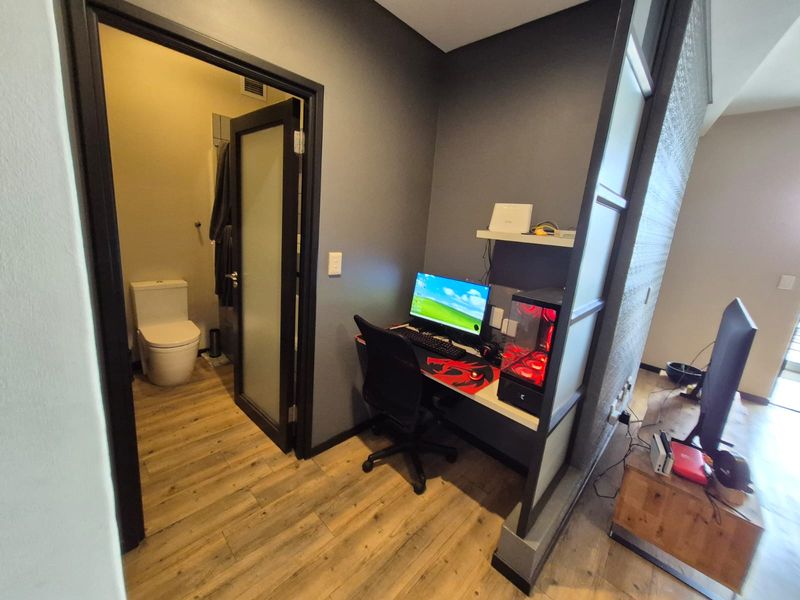 A compact home office nook featuring a built-in desk and floating shelf, adjacent to a modern bathroom with laminate flooring throughout.