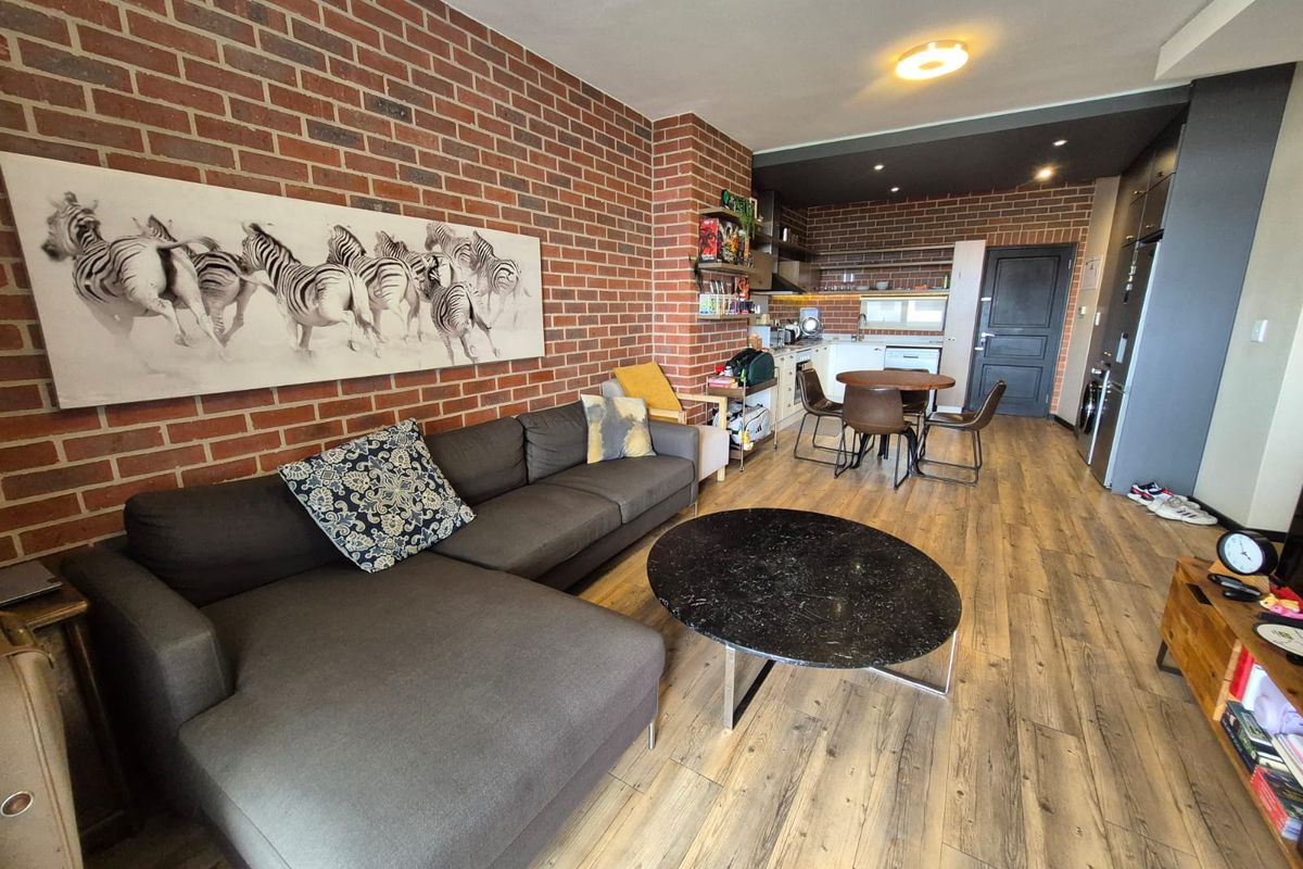 Open-plan living and kitchen area featuring a distinctive exposed brick feature wall, wood-look laminate flooring, and modern recessed lighting throughout the space.