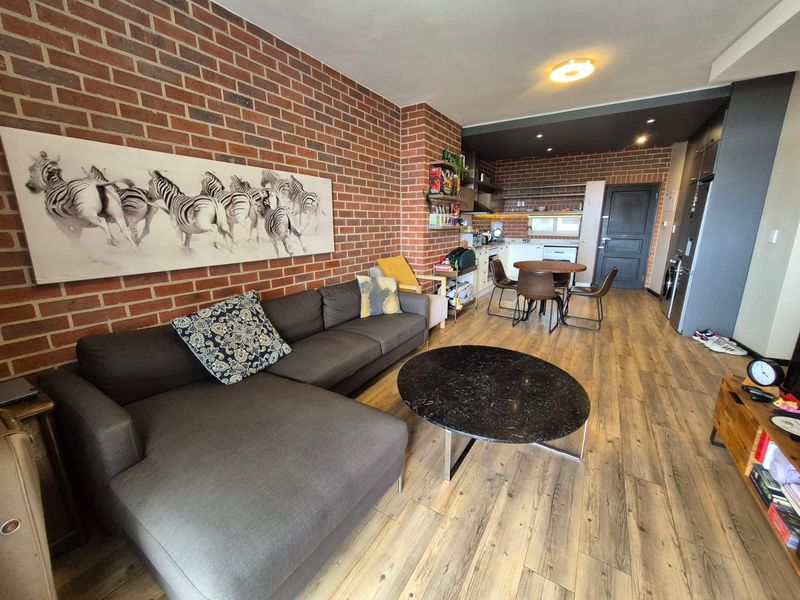 Open-plan living and kitchen area featuring a distinctive exposed brick feature wall, wood-look laminate flooring, and modern recessed lighting throughout the space.