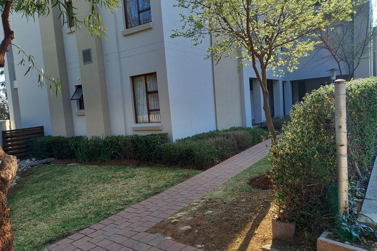 Exterior view of the apartment building featuring a paved walkway, landscaped garden area, and established trees alongside the building facade.