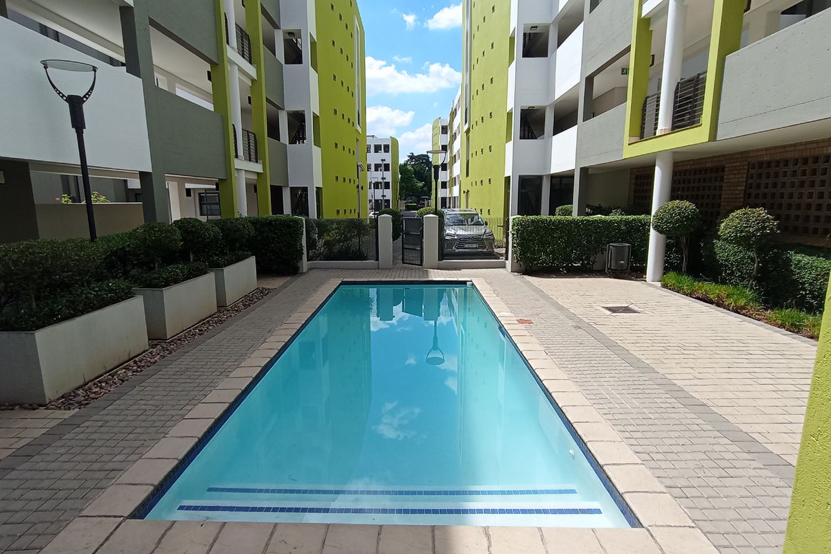 A well-maintained communal swimming pool area situated between apartment blocks, featuring paved surrounds and landscaped garden beds.