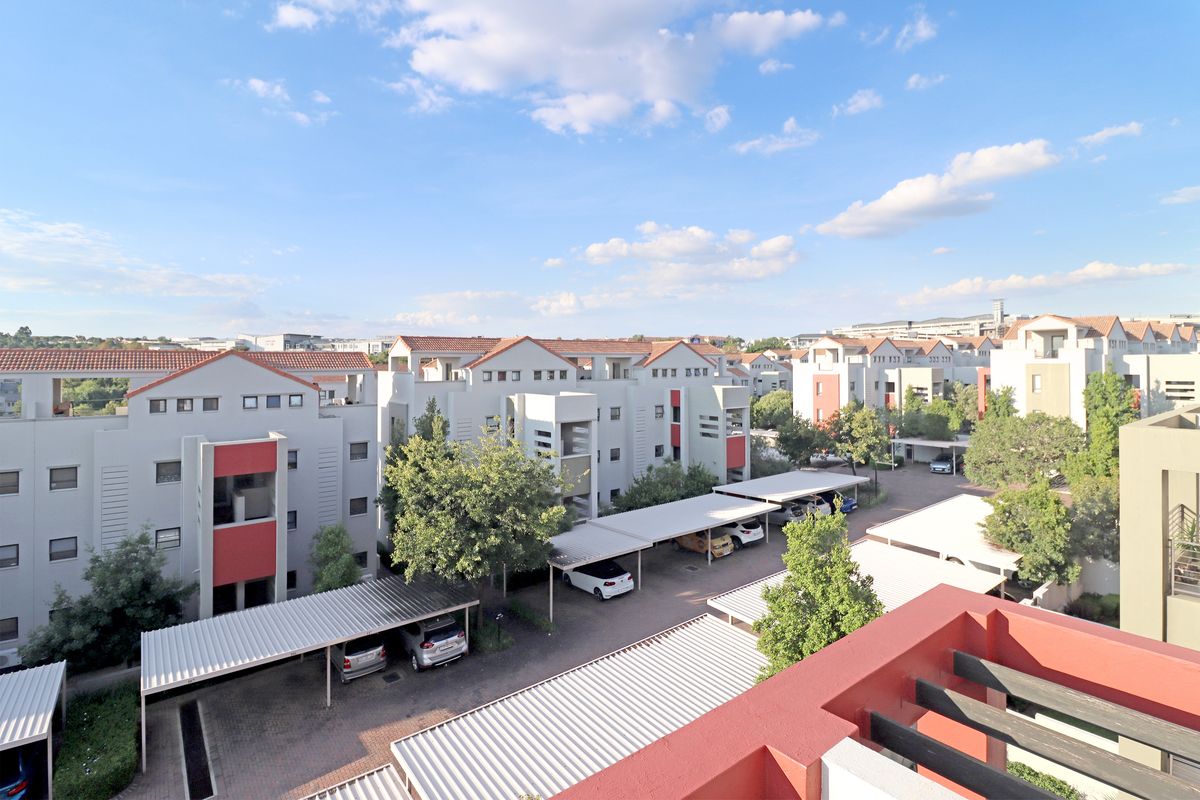 An elevated aerial view of the apartment complex, showcasing the residential buildings, paved parking areas with carports, and surrounding landscaped grounds.
