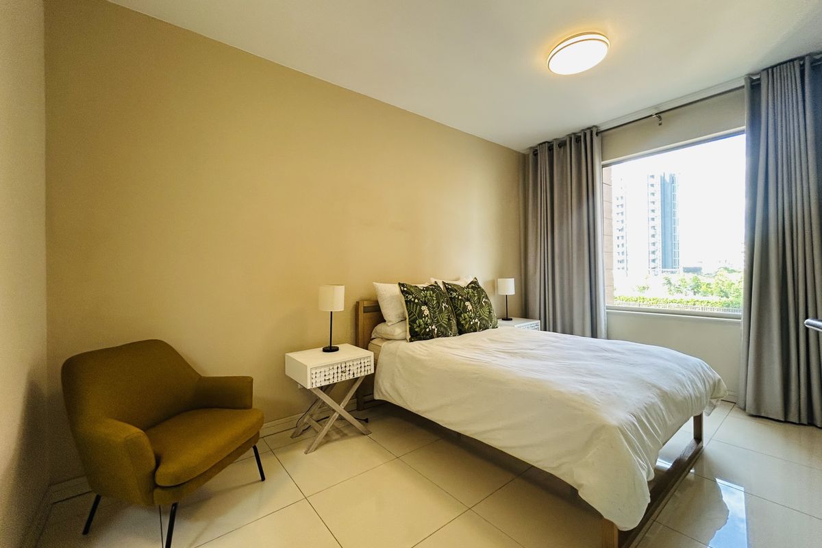 A well-lit bedroom featuring large windows with city views, neutral wall tones, and polished floor tiles.