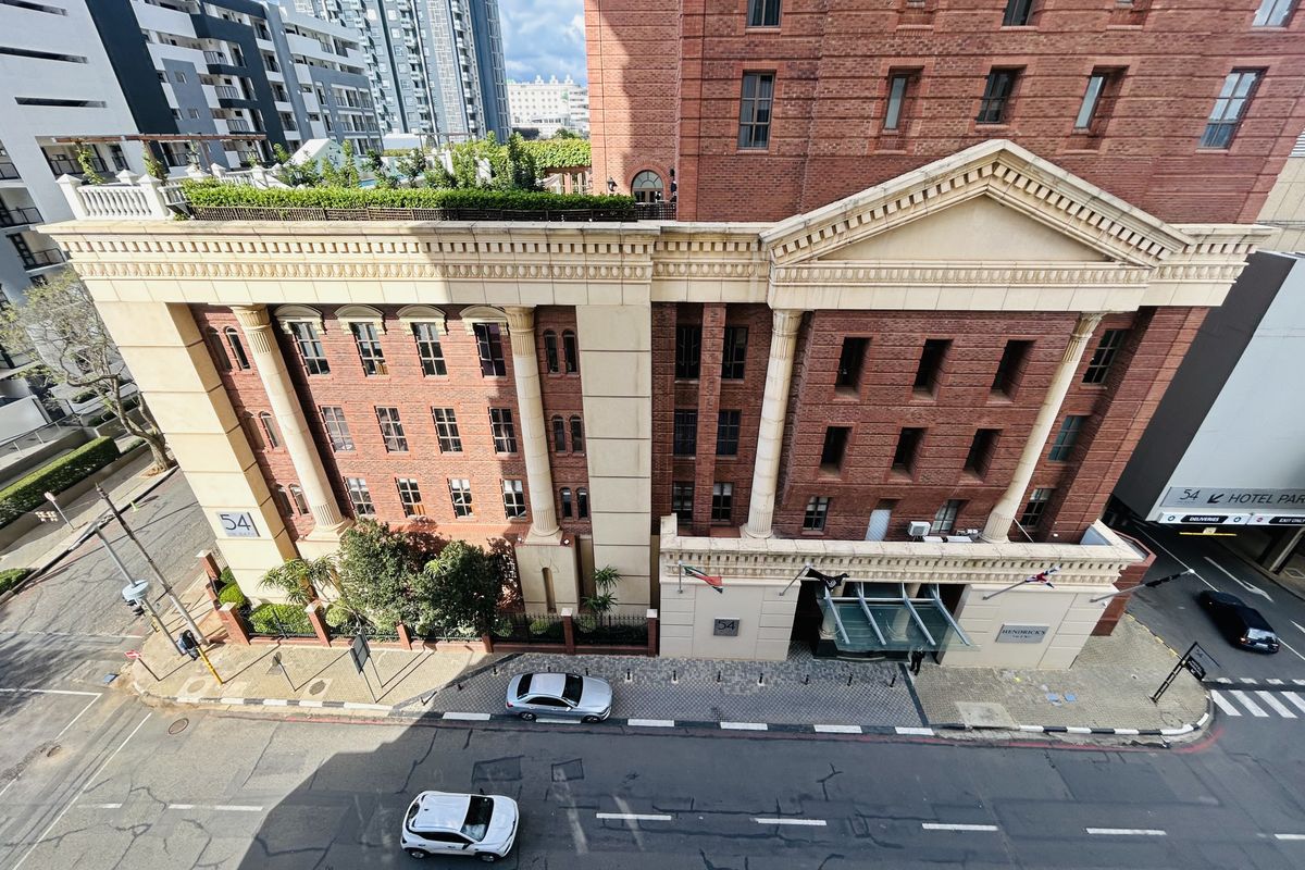 An elevated aerial view of a classic red-brick apartment building featuring architectural columns, a glass entrance canopy, and a rooftop garden terrace.