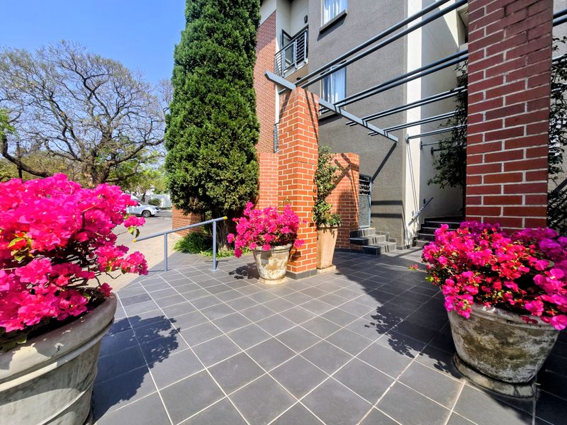 A well-maintained apartment building entrance featuring a tiled patio, brick architectural pillars, a metal pergola structure, and concrete access stairs.