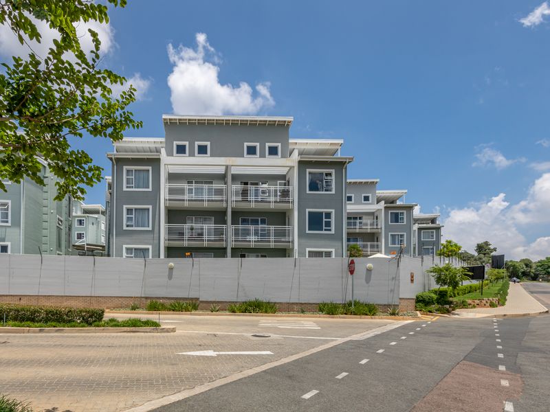 A clear street-level view of a modern apartment complex featuring grey facades, multiple balconies, and a secure perimeter wall.